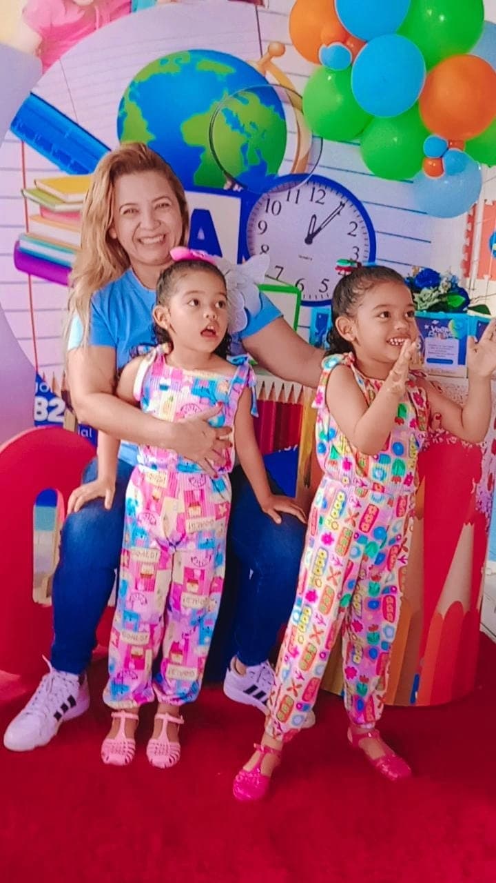 Educadora com duas alunas da Educação Infantil em comemoração de volta às aulas, com balões coloridos ao fundo