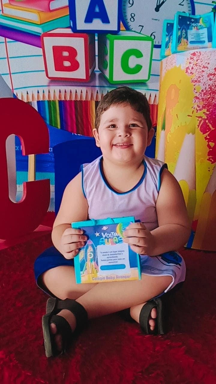 Menino sorridente segurando cartão de boas-vindas de volta às aulas, ao lado de blocos educativos com as letras A, B e C, no Colégio Baby Avançar