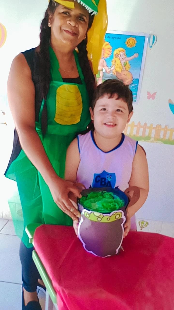 Educadora fantasiada de Curupira ao lado de aluno sorridente durante atividade do Dia do Folclore