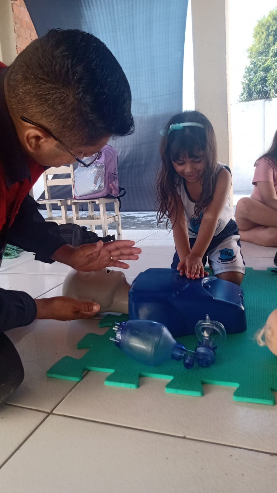 Criança praticando manobra de primeiros socorros em manequim de treino, orientada por instrutor