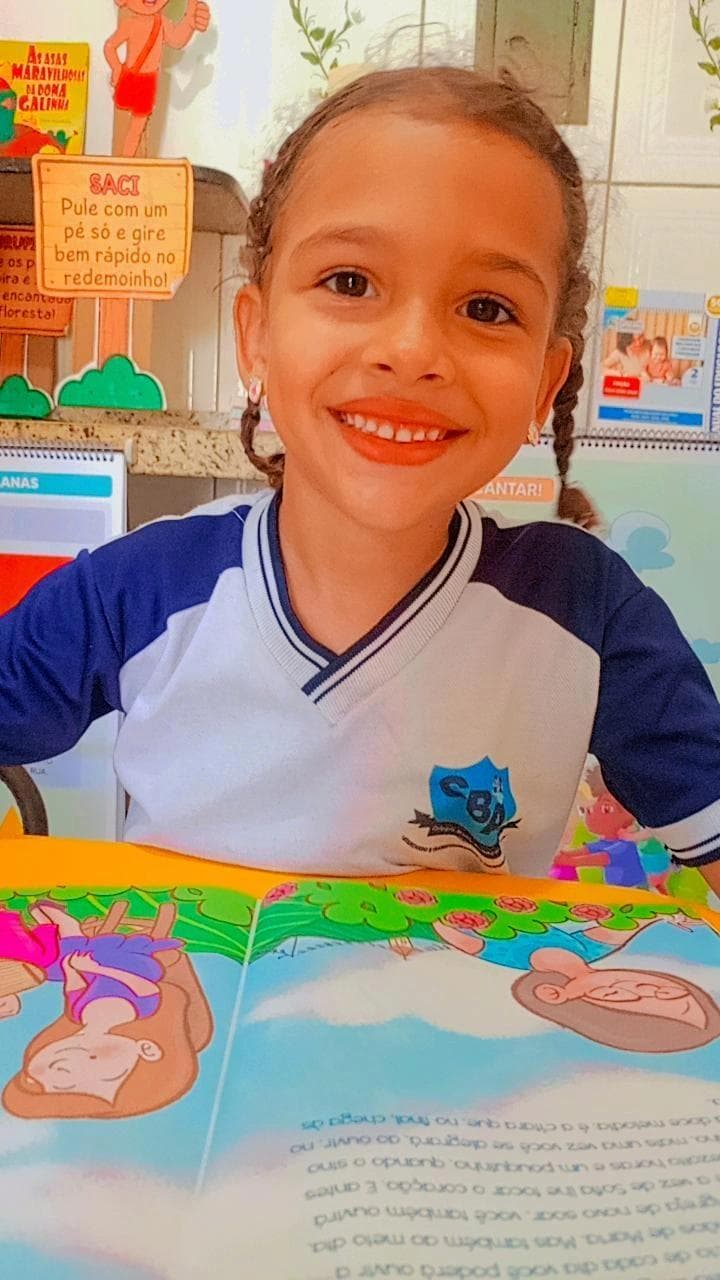 Aluna do Colégio Baby Avançar sorrindo com o uniforme da escola, lendo um livro infantil em sala de aula